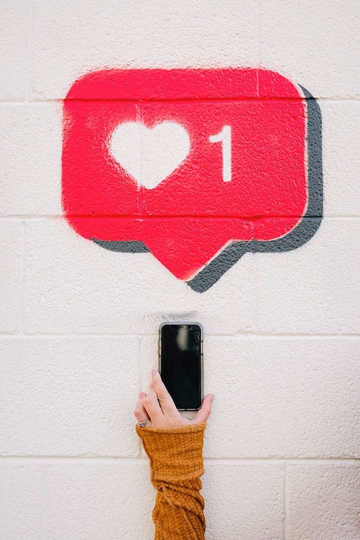 Photo of a hand holding a cell phone in front of a cinder block wall with a speech bubble above it with a hear and #1 inside it to represent the press release section of the site.